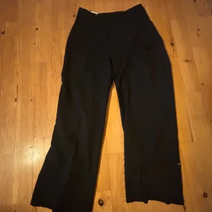 Svarta kostymbyxor från H&M - Svarta kostymbyxor från H&M med hög midja och raka, vida ben. Klassisk och stilren modell som passar till många olika tillfällen. Byxorna har diskreta fickor och är tillverkade i ett mjukt material.