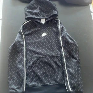 Svart hoodie från Nike med mönster - Säljer en svart hoodie från Nike med vitt logotryck på bröstet och diskret mönster över hela tröjan. Tröjan har huva, magficka och vita detaljer längs ärmarna. Perfekt för en sportig och avslappnad stil.