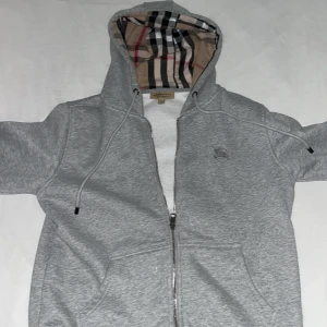 Grå hoodie med dragkedja från Burberry - Säljer en grå hoodie från Burberry med klassiskt rutigt foder i huvan. Jackan har dragkedja framtill, två fickor och broderad logga på bröstet. Perfekt för dig som gillar stilrena och exklusiva plagg.