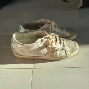 Golden Goose skor  - Säljer ett par vita sneakers från Golden Goose med glittrig stjärndetalj på sidan och beige snörning. Skorna har rund tå, platt sula och silverdetaljer vid hälen. Perfekta för dig som gillar en trendig och avslappnad stil.
