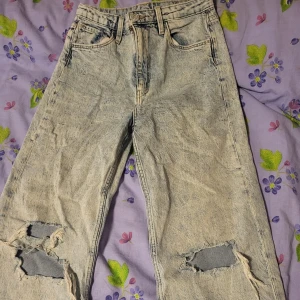 Ljusblå slitna jeans med vida ben - Säljer ett par ljusblå jeans med vida ben och stora slitningar vid knäna. Jeansen har klassisk femficksdesign och hög  midja. Perfekta för en avslappnad och trendig stil.