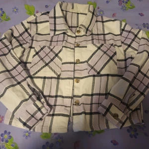 Rutig overshirt i beige och lila - Säljer en trendig overshirt i beige och lila med svart rutmönster. Jackan har två bröstfickor, knappar framtill och klassisk krage. Perfekt att slänga över en hoodie eller t-shirt för en avslappnad look.