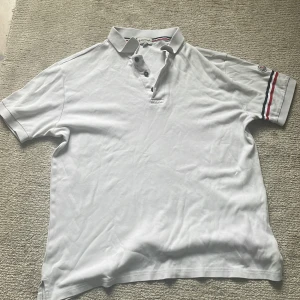 Vit piké från Moncler med röd och blå detalj - Vit pikétröja från Moncler med klassisk krage och knappar framtill. Ärmarna har röd och blå rand samt Moncler-logga på vänster ärm. Perfekt för en stilren och sportig look. 