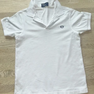Vit piké från Fred Perry - Klassisk vit pikétröja från Fred Perry i slim fit-modell. Tröjan har korta ärmar, krage med knappar och den ikoniska broderade loggan på bröstet. Perfekt för en stilren och sportig look. 