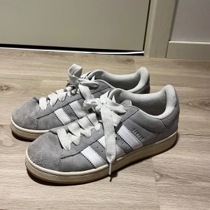 Adidas Campus grå sneakers - Säljer ett par Adidas Campus sneakers i grå mocka med vita detaljer och klassiska tre ränder på sidorna. Skorna har vit sula, rund tå och snörning. Perfekta för en stilren och avslappnad look. Skorna är i fint skick och har itne använts så mycket.