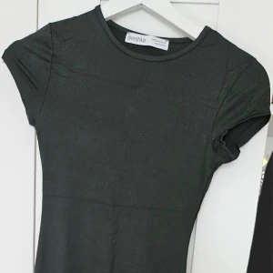 Grön ish långklänning från Bershka - En enkel och stilren grön långklänning från Bershka med rund halsringning och korta ärmar. Klänningen har en tight passform och är tillverkad i ett mjukt, stretchigt material som sitter snyggt på kroppen.