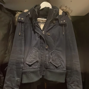 Mörkblå jacka med fuskpäls från Abercrombie & Fitch - Snygg mörkblå jacka med huva och fuskpäls från Abercrombie & Fitch. Jackan har dragkedja, knappar och ribbade muddar. Perfekt för kyligare dagar med sin varma huva och trendiga look. Dragkedjan är lite sönder så de kan va svårt att stänga men man måste vbara knepa lite men dragkedjan går att byta för typ 50kr hos skräddare.