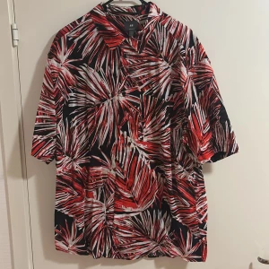 Röd och svart mönstrad skjorta från H&M - Säljer en färgstark skjorta från H&M med rött, vitt och svart palmbladsmönster. Skjortan har korta ärmar och avslappnad passform, perfekt för dig som gillar att sticka ut. Knappar framtill och klassisk krage.