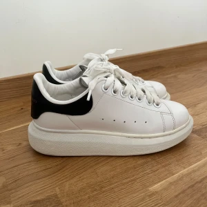 Vita sneakers från Alexander McQueen - Säljer ett par klassiska vita sneakers från Alexander McQueen med svart hälparti och tjock vit sula. Skorna har snörning och rund tå, samt diskret logga bak på hälen. 