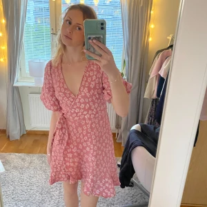 Rosa blommig omlottklänning från Pieces - Söt rosa omlottklänning från Pieces med blommigt mönster och volangdetaljer. Klänningen har kort ärm, v-ringning och knyt i midjan. Perfekt för sommarens alla tillfällen!