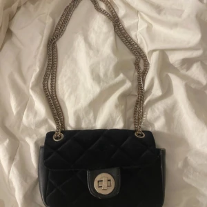 Svart quiltad axelväska från BikBok - Säljer en svart quiltad axelväska från BikBok med silverfärgad kedja och vridlås framtill. Väskan har en klassisk design och är perfekt för att lyfta vilken outfit som helst. Rymmer det viktigaste och är enkel att bära över axeln.