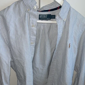 Blårandig skjorta från Polo Ralph Lauren - Klassisk blårandig skjorta från Polo Ralph Lauren i slim fit-modell. Skjortan har knappar framtill, button-down krage och den ikoniska broderade loggan på bröstet. Perfekt för en stilren och tidlös look.