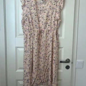 Blommig rosa klänning med volangdetaljer - Söt rosa klänning med blommigt mönster, volangdetaljer vid axlarna och knappar framtill. Klänningen har v-ringning, knytband i midjan och är ärmlös. Perfekt för varma dagar och somriga tillfällen.