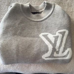 Grå stickad tröja från Louis Vuitton - Snygg grå stickad tröja från Louis Vuitton med stort vitt LV-logo på bröstet. Tröjan har rund halsringning och långa ärmar. Perfekt för dig som gillar exklusiva och stilrena plagg.