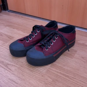 Vinröda sneakers med svarta detaljer - Säljer ett par vinröda sneakers med svarta snören och svarta detaljer runt sula och tå. Skorna har rund tå, platt sula och coolt mönster under sulan. Perfekta för dig som vill ha en stilren men ändå unik look.