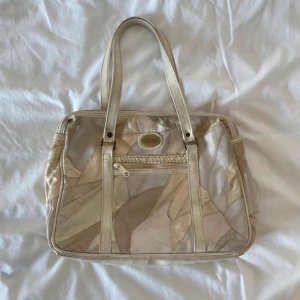 Beige handväska i patchwork från Tazzio - Säljer en beige handväska från Tazzio med patchwork-design i olika nyanser av beige och kräm. Väskan har dubbla handtag, dragkedja upptill och en liten ficka med dragkedja framtill. Perfekt storlek för vardagens alla prylar.