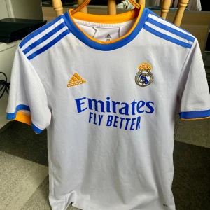 Real Madrid hemmatröja 2021/22 från Adidas - Säljer en vit Real Madrid hemmatröja från Adidas med blå och gula detaljer. Tröjan har korta ärmar, klubbmärke på bröstet och Adidas-logga. Klassiska blå ränder på axlarna och sponsortryck framtill.