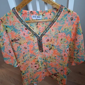 Blommig blus från Vero Moda - Säljer en färgglad blommig blus från Vero Moda i storlek XS. Blusen har v-ringning med dekorativa pärldetaljer och korta ärmar. Materialet är lätt och luftigt, perfekt för varmare dagar. Mönstret går i orange, turkos, gul och svart.