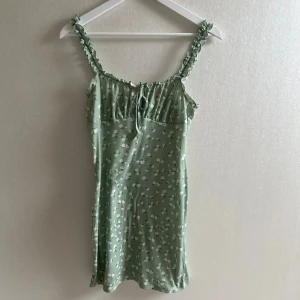 Ljusgrön kort blommig klänning  - Supersöt ljusgrön klänning med små vita blommor, volangkant upptill och knyt framtill. Axelbanden är rynkade och klänningen har en mjuk, ribbad struktur. Perfekt för varma dagar!🩷