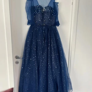 Blå glittrig balklänning med stjärnmönster - Magisk blå långklänning med glittrigt stjärnmönster, pärldetaljer och broderade blommor i midjan. Klänningen har mesh-tyg upptill, snörning i ryggen och är ärmlös. Perfekt för bal eller festliga tillfällen.