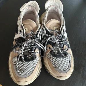 Beige och svarta sneakers från Bronx - Säljer ett par chunky sneakers från Bronx i beige och svart med meshdetaljer och mönstrade snören. Skorna har en robust sula och sportig design, perfekta för en trendig vardagslook.