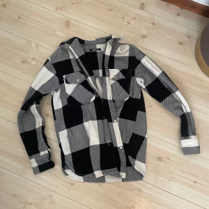 Svartvit rutig overshirt från Gina  - Säljer en oversized svartvit rutig skjorta med stora rutor och två bröstfickor med lock. Skjortan har långa ärmar och knäppning framtill. Perfekt att slänga över en t-shirt för en avslappnad look.