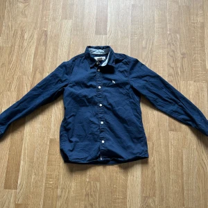 Mörkblå skjorta från L.O.G.G. - Säljer en klassisk mörkblå skjorta från L.O.G.G. med långa ärmar och vita knappar. Skjortan har en liten broderad logga på bröstet och är tillverkad i bomull. Perfekt för en stilren look.