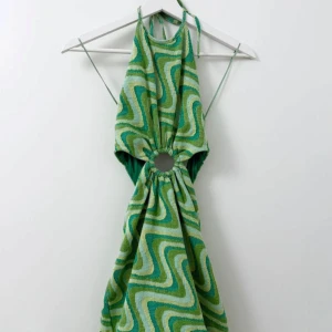 Grön mönstrad klänning från Zara - Retroinspirerad grön klänning från Zara med vågigt mönster i olika gröna och gula nyanser. Klänningen har halterneck, öppen rygg och en dekorativ ringdetalj framtill. Perfekt för sommarens alla tillfällen.
