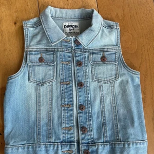 Ljusblå jeansväst från OshKosh B’gosh - Säljer en klassisk ljusblå jeansväst från OshKosh B’gosh med bruna knappar och två bröstfickor med lock. Storlek barn 8 år.