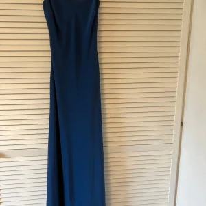 Blå långklänning med smala axelband - Elegant blå långklänning med smala axelband och dekorativ kedjedetalj i ryggen. Klänningen har en rak, slimmad passform och är perfekt för festliga tillfällen. Materialet har en lätt glans och ger ett stilrent intryck. Obs - finns fläckar, kanske går bort i tvätt.