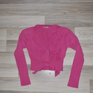 Rosa croppad kofta från NONO - Säljer en rosa croppad kofta från NONO med v-ringning och knappar framtill. Tröjan har långa ärmar och ribbad nederkant. Perfekt för dig som vill ha en färgglad och trendig look.