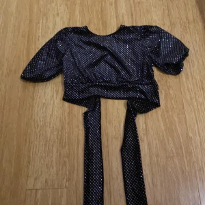 Svart glittrig croppad topp från Divided - Säljer en svart glittrig croppad topp från Divided med knytband framtill. Toppen har korta puffärmar och är täckt av små silvriga paljetter som ger en festlig look. Perfekt för att sticka ut på nästa party!