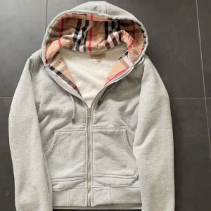 Grå hoodie med dragkedja från Burberry - Säljer en grå hoodie från Burberry med klassiskt rutigt foder i huvan. Tröjan har dragkedja framtill, två fickor och justerbar huva med snören. Perfekt för en avslappnad och stilren look.