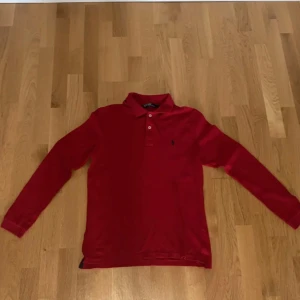 Röd långärmad pikétröja från Ralph Lauren - Säljer en klassisk röd långärmad pikétröja från Ralph Lauren med krage och knappar framtill. Tröjan har den ikoniska broderade loggan på bröstet och är tillverkad i bomull. Perfekt för en stilren och avslappnad look.