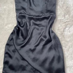 Svart satinklänning från BikBok - Säljer en svart, figurnära satinklänning från BikBok i storlek XS. Klänningen har en elegant, slät yta och är axelbandslös med en kort längd. Perfekt för dig som vill ha en stilren och klassisk look.