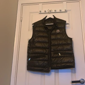 Oliv grön dunväst från Moncler - Säljer en svart dunväst från Moncler med klassisk logotyp på bröstet. Västen har dragkedja framtill och två fickor med dragkedja. Perfekt för lager-på-lager och stilren look. Har äkthets bevis