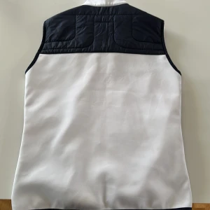 Marinblå och vit quiltad väst från J.Lindeberg - Snygg marinblå och vit quiltad väst från J.Lindeberg med dragkedja framtill och två fickor med vita dragkedjor. Västen är andvänd fåtal gånger för den är för liten. Storlek S. 