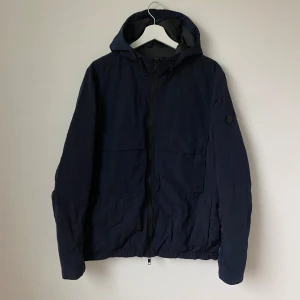 Mörkblå vindjacka med huva från J.Lindeberg - Snygg mörkblå vindjacka från J.Lindeberg med huva och dragkedja framtill. Jackan har en minimalistisk design och är tillverkad i ett lätt syntetmaterial, perfekt för blåsiga dagar. Justerbar nederkant och diskret logga på ärmen. Modell: Bass Nickel Memo - Artnr: Z246