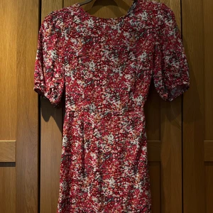 Röd blommig klänning med puffärm - Säljer en röd blommig klänning med vita och rosa inslag. Klänningen har korta puffärmar, rund halsringning och en öppen detalj i ryggen. Modellen är figurnära upptill och har en feminin siluett. Perfekt för sommarens tillfällen!