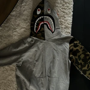 Grå hoodie med kamouflagemönster av Bathing Ape - Säljer en OANVÄND grå hoodie från BAPE med kamouflagemönstrade ärmar och huva. Huvan har hajansikte med tänder och ögon, samt broderade bokstäver och tigerdetalj. Dragkedja hela vägen upp och fickor framtill. Pris kan diskuteras. 