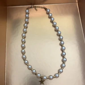 Pärlhalsband med stjärnhänge - Säljer ett elegant halsband med vita pärlor och ett guldigt stjärnhänge. Halsbandet har justerbart lås och passar perfekt för att ge din outfit en extra touch av stil.