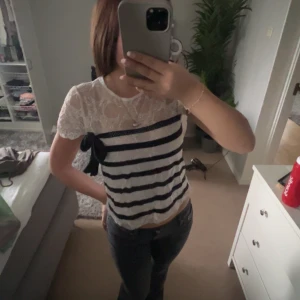 Vit och marinblå randig blus med spets - Säljer en söt blus med vit spets upptill och marinblå horisontella ränder nedtill. Blusen har en dekorativ rosett på ena sidan som du kan knyta själv. Perfekt för dig som gillar detaljer och en feminin stil.