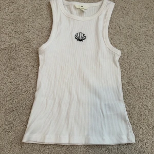 Vit ribbad linnetopp med snäckbrodyr från H&M - Säljer en vit ribbad linnetopp från H&M med en liten svart snäckbrodyr framtill. Toppen är ärmlös och har en klassisk rund halsringning. Perfekt för varma dagar eller som lager under andra plagg.