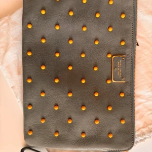 Grå clutch från Marc Jacobs med nitar - Säljer en grå clutch från Marc Jacobs med orangea nitar och guldfärgad logotypplatta. Väskan är i skinn och har dragkedja upptill. Perfekt accessoar för att lyfta din outfit.