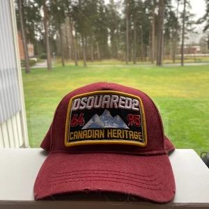 Vinröd keps från Dsquared2 med broderad patch - Vinröd keps från Dsquared2 med stor broderad patch framtill där det står 'Canadian Heritage' och bergmotiv. Justerbar rem baktill i samma färg. Kepsen har böjd skärm och tydliga broderade detaljer.