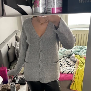 Grå kofta med v-ringning - Mysig grå kofta med v-ringning och knappar framtill. Tröjan har långa ärmar och två fickor framtill. Perfekt att slänga på sig över en t-shirt eller linne för en avslappnad look.