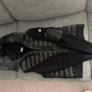 Svart quiltad väst med huva från Moncler - Säljer en svart quiltad väst från Moncler med huva och dragkedja. Västen har stickade partier på sidorna och längs ärmhålen för extra komfort och stil. Perfekt för lager-på-lager och snygg Moncler-logga på sidan. Den är i mycket bra skick och inte använd så mycket. Pris kan diskuteras vid snabb affär! 