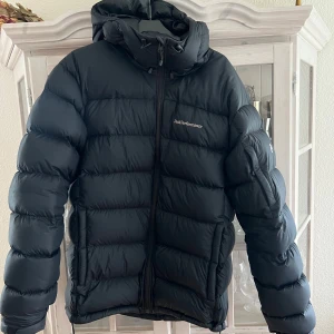 Svart dunjacka från Peak Performance - Säljer en svart dunjacka från Peak Performance med huva och dragkedja. Jackan är vadderad och har Pertex Quantum-material för extra hållbarhet och lätthet. Perfekt för kyliga dagar.
