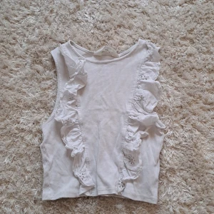 Vit ribbad linnetopp med volang från H&M - Säljer en söt vit ribbad linnetopp från H&M med volangdetaljer framtill. Toppen är ärmlös och har en mjuk, stretchig känsla. Perfekt för varma dagar och enkel att matcha med olika outfits.