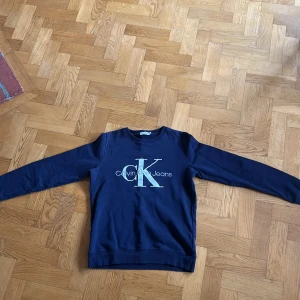 Calvin Klein Hoodie - Säljer en mörkblå sweatshirt från Calvin Klein Jeans med stort CK-tryck på bröstet. Tröjan har rund halsringning och långa ärmar. Perfekt för en stilren och avslappnad look. Kontakta vid minsta fråga 🤗